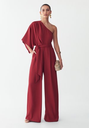 Donna che indossa una tuta bordeaux monospalla con gambe larghe, tiene una clutch intrecciata, accessoriata con orecchini e braccialetto oro.