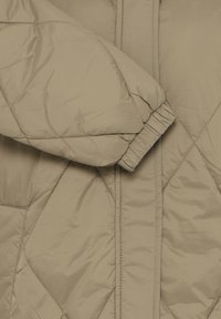 Beige gesteppte Jacke mit ärmellosem Gummiabschluss, die auf einem passenden gesteppte Stoffhintergrund ruht.