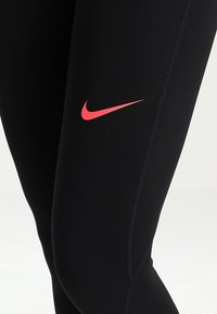 Czarne sportowe legginsy o gładkiej fakturze, z kontrastującym czerwonym logo Nike w kształcie łyżwy na lewej uda.