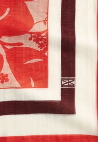 Tissu rouge et crème à motifs floraux, avec une bordure marron foncé et un petit motif géométrique dans un coin.