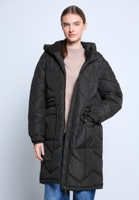 Giovane donna che indossa un lungo piumino trapuntato nero sopra un maglione beige e jeans blu, in piedi con una mano nella tasca.