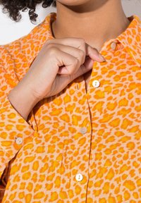 Košile s oranžovým leopardím vzorem a límečkem, malými bílými knoflíky a texturovanou tkaninou. Model si upravuje knoflík límečku.