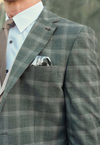 Blazer grigio a quadri con una finitura strutturata, dotato di una tasca sul petto con un fazzoletto a fantasia e una camicia dress di colore chiaro sotto.