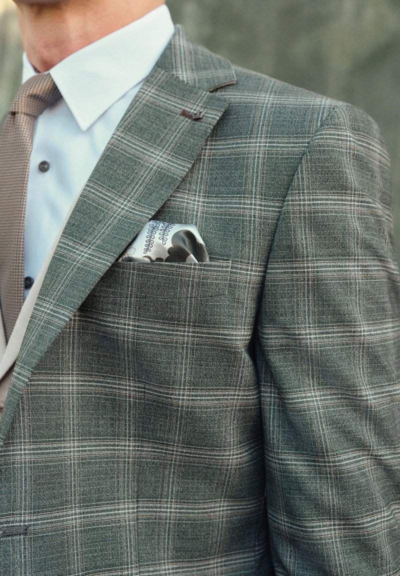 Blazer grigio a quadri con una finitura strutturata, dotato di una tasca sul petto con un fazzoletto a fantasia e una camicia dress di colore chiaro sotto.