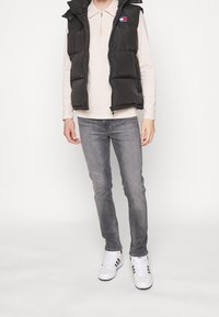 Schwarze, puffy Weste mit hohem Kragen, getragen über einem hellbeigen Langarmshirt. Graue Jeans und weiße Sneakers mit schwarzen Streifen runden den Look ab.