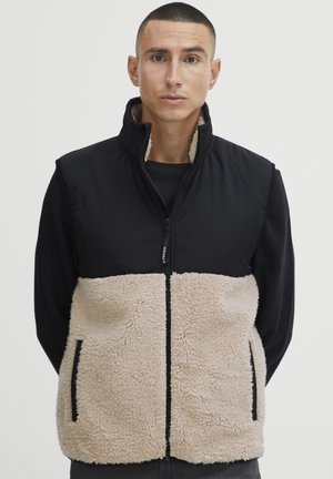 Black and beige sleeveless vest with a zip front. Upper section is smooth fabric; lower section features textured, fluffy material. Side pockets.