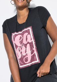 Mujer sonriente que viste una camiseta negra con un gráfico rectangular en rosa y blanco que dice "Perezosa como un domingo por la mañana" y pendientes de aro.