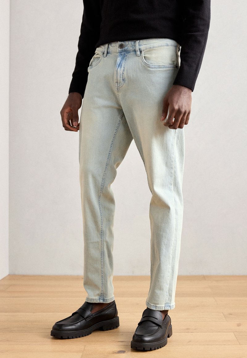 Light blue denim jeans featuring a straight-leg design, slight fading, and a five-pocket style. Paired with black slip-on shoes.