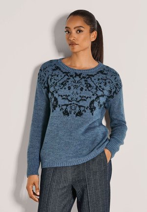 Frau mit dunklem Haar im Pferdeschwanz, die einen blauen Strickpullover mit schwarzem Blumenmuster und eine dunkelgraue, gestreifte Hose trägt, steht vor einer hellen Wand.