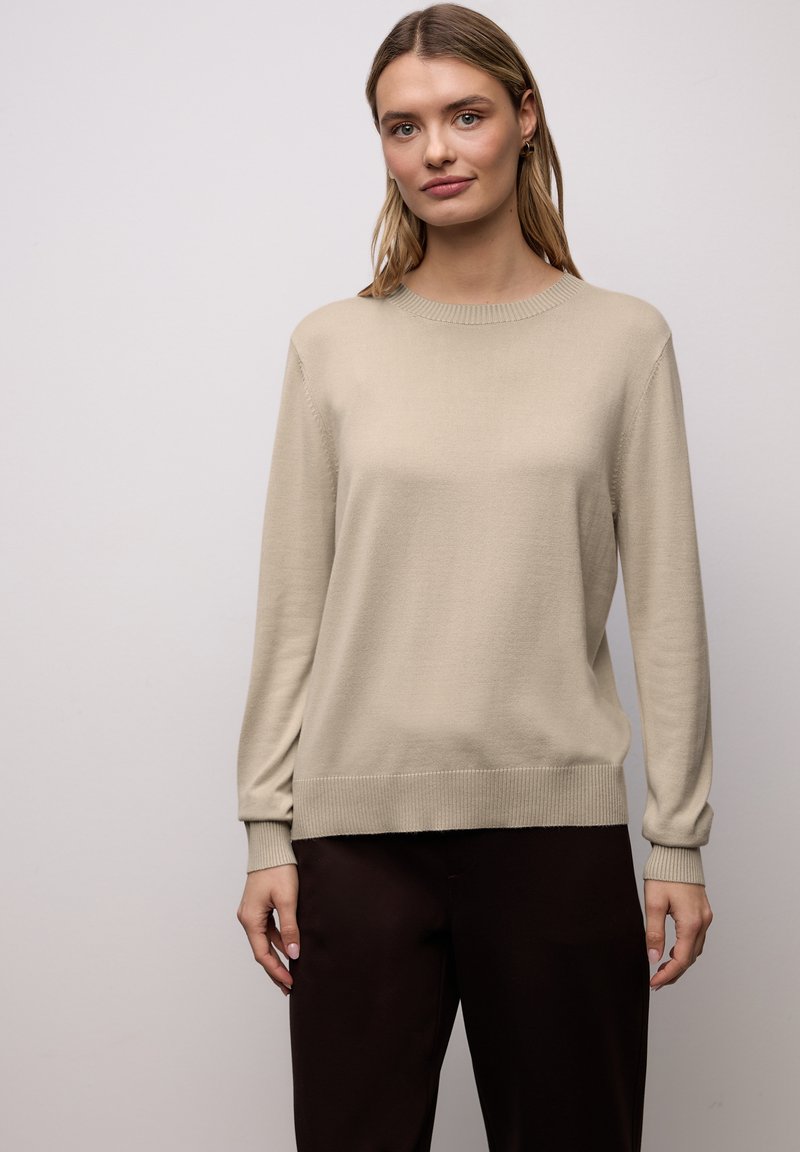 Pull beige à manches longues avec encolure ronde, poignets et ourlet côtelés, fabriqué en tissu tricoté doux. Associé à un pantalon brun foncé.