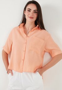 Short-sleeve, button-up shirt in light orange linen with a chest pocket, collar, and relaxed fit, paired with white trousers.