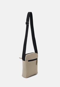 Beige crossover bag med glatt materiale, svart justerbar stropp, glidelåslomme foran og rektangulær form.