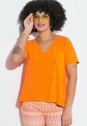 Kvinde med krøllet hår iført gule solbriller, skrigorange V-hals trøje, mønstrede bukser og guldbøjle, poserer mod hvid baggrund.