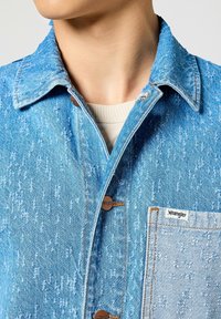 Giubbotto di jeans blu chiaro con una finitura texturizzata, bottoni marroni e una tasca applicata. Presenta un colletto e dettagli di cucitura a contrasto.