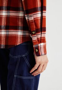 Camisa de franela a cuadros en rojo y blanco con puños abotonados, acompañada de pantalones cargo azul oscuro con costuras en contraste.