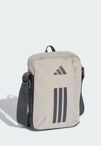 Prostokątna beżowa torba z ciemnymi akcentami, posiadająca zapinaną na zamek kieszeń, regulowany pasek oraz logo z trzema czarnymi paskami z przodu.