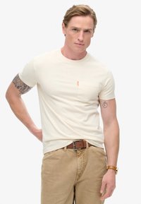 Cremefarvet t-shirt med korte ærmer og brystlomme samt orange accent. Bæres med beige bukser og et brunt bælte. Glat tekstur, tætsiddende design.