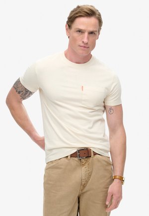 Cremefarvet t-shirt med korte ærmer og brystlomme samt orange accent. Bæres med beige bukser og et brunt bælte. Glat tekstur, tætsiddende design.