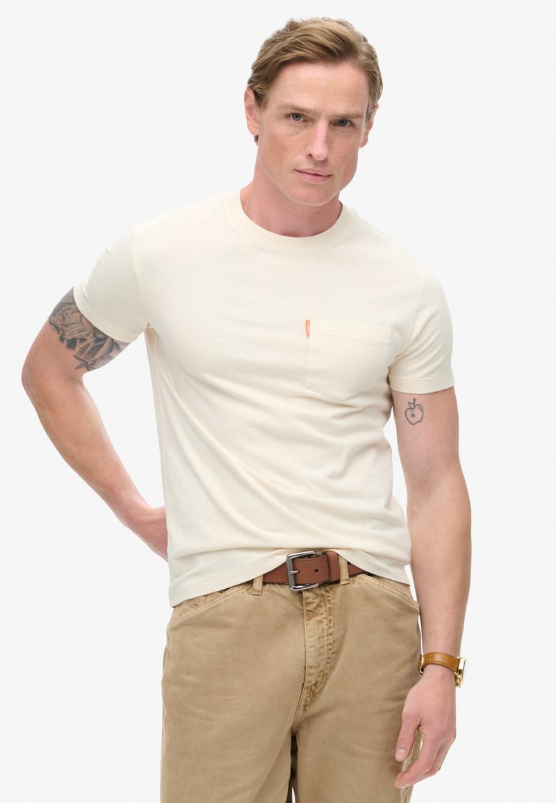 Cremefarvet t-shirt med korte ærmer og brystlomme samt orange accent. Bæres med beige bukser og et brunt bælte. Glat tekstur, tætsiddende design.