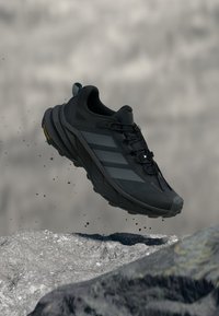 Chaussure de sport noire avec une tige texturée, des accents de design à trois bandes, et une semelle robuste, montrée en suspension au-dessus d'un terrain rocheux.