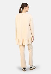 Beige tuniek met een gerimpelde zoom, lange mouwen en een keyhole-detail aan de achterkant, gecombineerd met bijpassende strakke broek en witte sneakers.