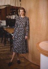 Bruine bloemen midi-jurk met witte accenten, lange mouwen, geplooide halslijn, geplooide rok, gecombineerd met donkere instappers. Achtergrond van houten panelen.