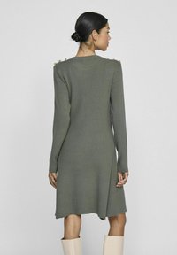 Robe tricotée verte à manches longues, texture côtelée, boutons sur les épaules, et coupe évasée, tombant mi-cuisse, assortie avec des bottes claires.
