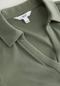 Olive green ribbed polo shirt with a collar, featuring a label sewn on the neckline, showcasing a soft texture and detailed stitching.