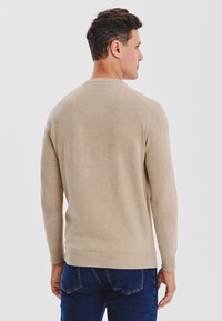 Beżowy sweter z długim rękawem i okrągłym dekoltem, wykonany z gładkiego materiału dzianinowego, z prążkowanymi mankietami i dołem, noszony z niebieskimi dżinsami.