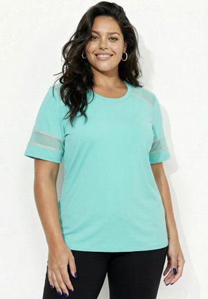 Smilende kvinde med langt mørkt hår iført en turkis kortærmet top med mesh-stribedetaljer og sorte bukser, stående foran en hvid baggrund.