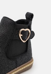 Černé kotníkové boty s texturovaným povrchem, zdobené zlatým akcentem ve tvaru srdce a elastickým bočním panelem. Hnědá podrážka s bílými stehy.