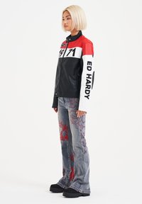 Black leather jacket with red and white accents, featuring "ED HARDY" and "1971" graphics. Gray flared jeans with red floral embroidery. Chunky black shoes.