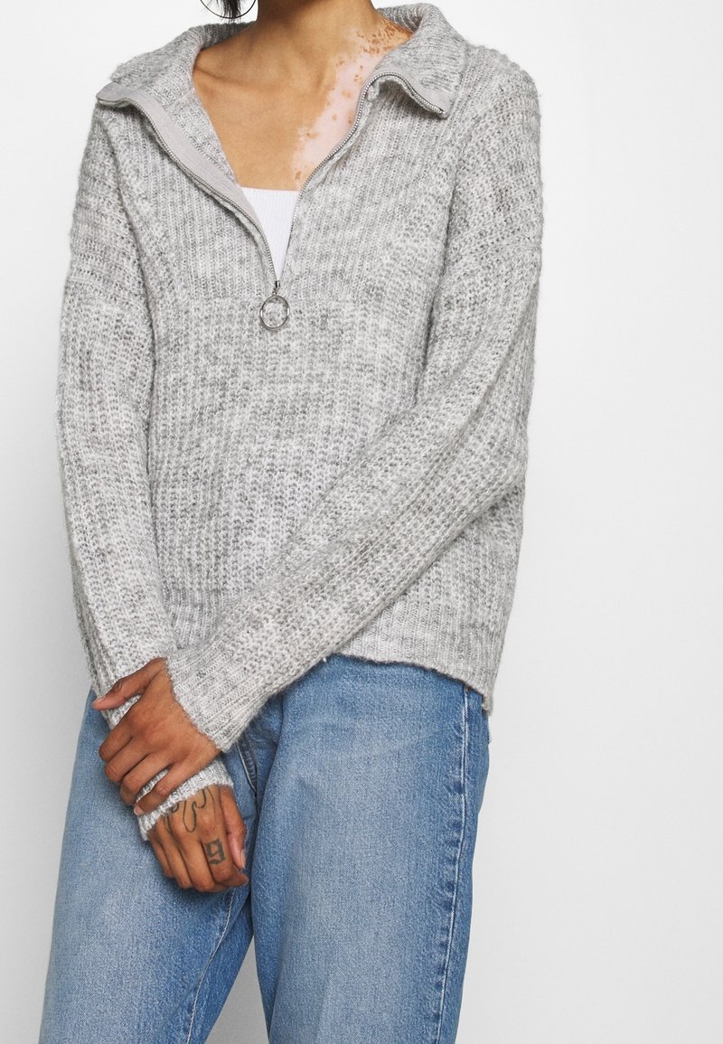 Personne portant un pull en maille gris clair avec un col demi-zip et un jean bleu, bras croisés, tatouages visibles sur la main gauche.