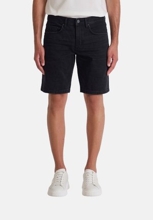 Hombre con pantalones cortos de mezclilla negros y zapatillas blancas, de pie con los brazos relajados a los lados sobre un fondo liso.