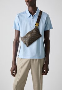 Pruun mustriga ristiõla kott kuldse riistvaraga, kantud taevasinise polo särgi peal, koos heledate beežide pükstega.