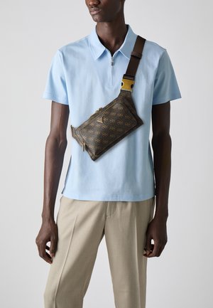 Hnedá vzorovaná crossbody taška so zlatým hardvérom, nosená cez svetlomodré polo tričko, v kombinácii so svetlobežovými nohavicami.