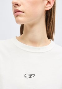 Κοντινό πλάνο του κάτω μέρους του προσώπου, του λαιμού και του ώμου ενός ατόμου που φοράει λευκό πουκάμισο με ένα μικρό μαύρο αφηρημένο λογότυπο στο στήθος.