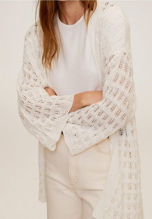 Cardigan blanc en tricot ouvert avec de larges manches, présentant un motif en dentelle délicat. Porté par-dessus un haut blanc uni, assorti à un pantalon de couleur claire.