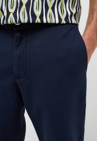 Granatowe bawełniane spodnie o gładkiej fakturze, z kieszeniami po bokach i prostym krojem. Pas zapinany jest na guzik.