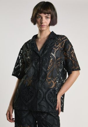 Młoda osoba z krótkimi brązowymi włosami ubrana w czarną koronkową bluzkę z krótkim rękawem i dopasowane spodnie, stojąca na tle jasnej, jednolitej ściany.