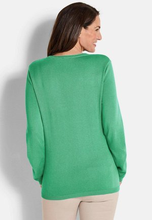 Femme aux cheveux bruns souriante, portant un pull vert à manches longues et un pantalon beige, de dos contre un fond blanc uni.
