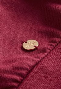 Burgundská látka s hladkým povrchem, zdobená zlatým kovovým knoflíkem s reliéfním květinovým vzorem, přišitým červenou nití.
