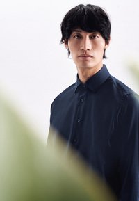 Camicia a button-up di un blu navy scuro con colletto, caratterizzata da una texture liscia e bottoni visibili, parzialmente oscurata da fogliame verde.