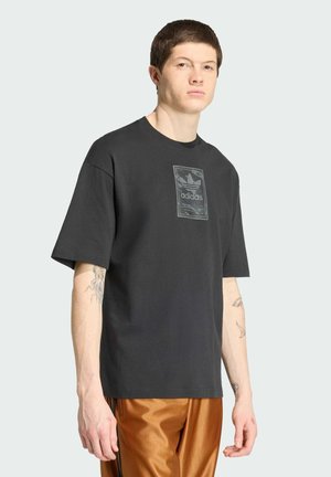 Junger Mann mit kurzen dunklen Haaren, der ein schwarzes Adidas-T-Shirt mit Logo und braune Hose trägt, steht vor einem hellen einfarbigen Hintergrund.
