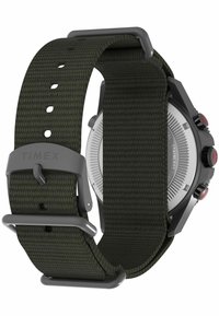 Reloj Timex con una correa de tela trenzada de color verde oscuro, caja de acero inoxidable y acentos rojos en los pulsadores. Carátula redonda con textura visible.