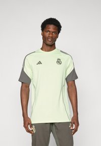 T-shirt de sport vert clair avec des manches courtes grises, présentant le logo du Real Madrid et la marque Adidas. Fabriqué en tissu lisse, avec un col rond.