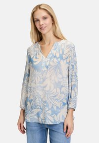 Blusa azzurro chiaro realizzata in tessuto morbido e semitrasparente con un motivo floreale crema, caratterizzata da un colletto a V e maniche tre quarti.