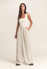 Beige viddebukser i let stof, med høj talje, plisseret front og kombineret med en tætsiddende hvid top.