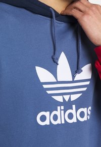 Hooded sweatshirt in blue fabric with a textured finish. Features a large white Adidas logo and contrasting red accents on the sleeves.