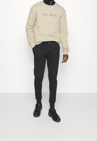 Hombre con sudadera beige que lleva el texto "LES DEUX", pantalones joggers negros ajustados y zapatos negros, de pie contra un fondo blanco liso.